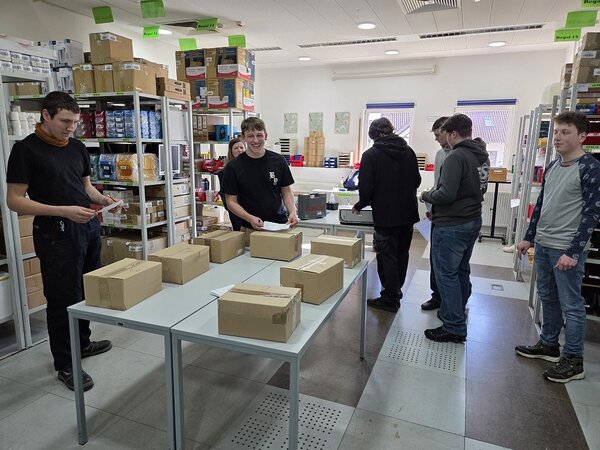 In einem hellen Raum mit Regalen voller Pakete steht in der Mitte ein Tisch mit mehreren Päckchen, drumherum stehen mehrere junge Menschen mit Zetteln in den Händen.