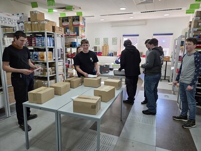 In einem hellen Raum mit Regalen voller Pakete steht in der Mitte ein Tisch mit mehreren Päckchen, drumherum stehen mehrere junge Menschen mit Zetteln in den Händen.