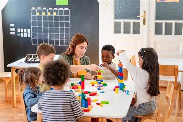 Eine Gruppe Kindergartenkinder baut zusammen mit einer Betreuerin Türme aus bunten Holzklötzen; im Hintergrund ist eine Tafel zu sehen.