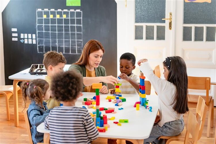 Eine Gruppe Kindergartenkinder baut zusammen mit einer Betreuerin Türme aus bunten Holzklötzen; im Hintergrund ist eine Tafel zu sehen.