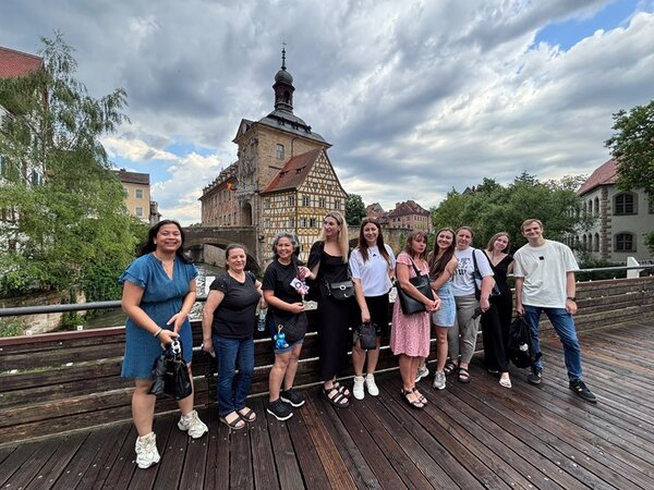Die Gruppe der Teilnehmenden des Orientierungskurses steht auf einer Holzbrücke vor einem historischen Fachwerkgebäude mit Turm und Uhr. Im Hintergrund sind weitere Gebäude mit roten Dächern und Bäume zu sehen. Der Himmel ist bewölkt. Die Personen tragen sommerliche Kleidung und blicken in Richtung Kamera.