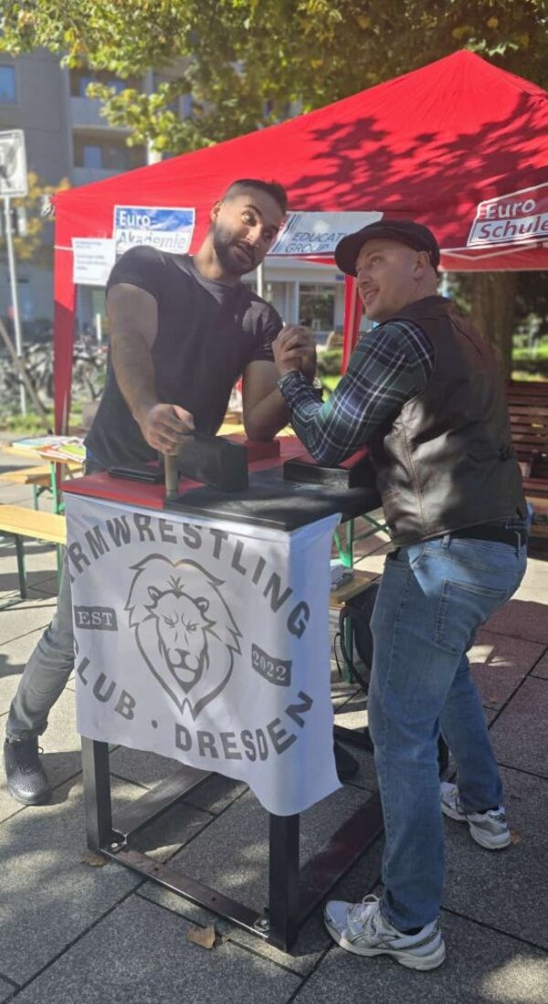 Zwei Männer armwresteln an einem kleinen Tisch mit dem Banner „Armwrestling Club Dresden est. 2022", dahinter das rote Zelt der Euro-Schulen auf dem gepflasterten Festplatz.
