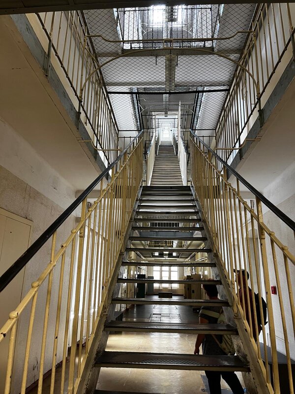 Blick in das hohe, mehrstöckige Treppenhaus eines Gefängnisses mit Gittern und engen Fluren. Zwei Personen stehen am unteren Treppenabsatz und blicken hinauf.