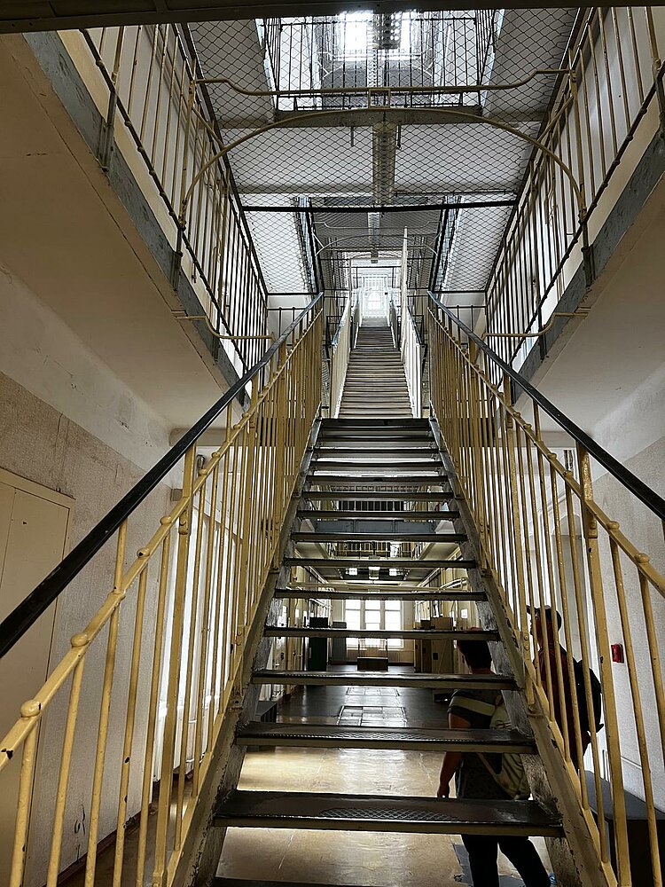 Exkursion zur Gedenkstätte Bautzen Blick in das hohe, mehrstöckige Treppenhaus eines Gefängnisses mit Gittern und engen Fluren. Zwei Personen stehen am unteren Treppenabsatz und blicken hinauf.