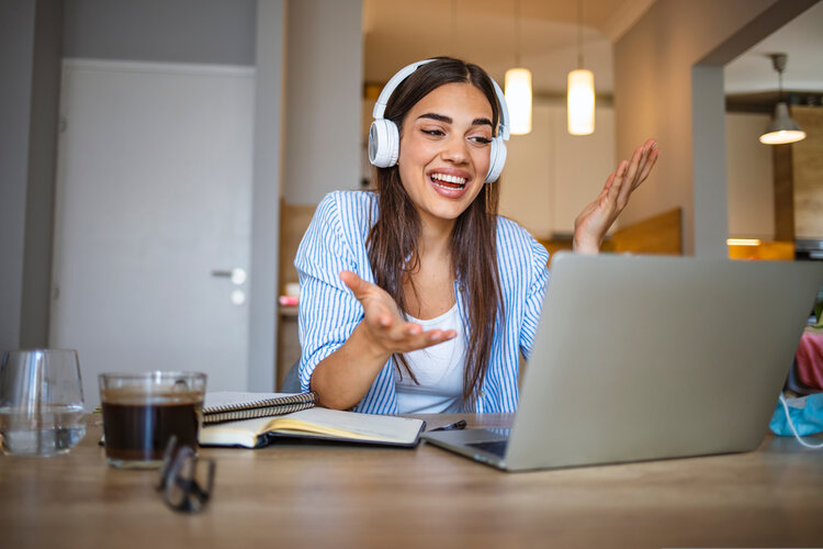Eine Frau mit Kopfhörern sitzt vor ihrem Laptop und guckt freundlich in den Bildschirm. Sie gestikuliert mit den Händen.