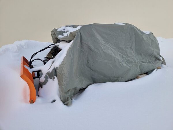Die orangefarbene Schneeschaufel des Schneeräumfahrzeugs ist zu sehen, der Rest ist von einer grauen Plane bedeckt. Die Schneeschaufel und die Plane sind von einer dicken Schneeschicht umgeben. Im Hintergrund erstreckt sich eine helle Gebäudewand