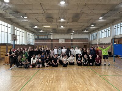 Gruppe aus Teilnehmenden des Volleyballturniers posiert in der Sporthalle, einige halten Volleybälle, vorne springt eine Person im neongrünen Shirt begeistert in die Luft.