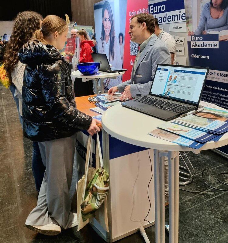 Zwei junge Frauen stehen am Stand der Euro Akademie auf dem Karrieretag Oldenburg 2025. Ein Mitarbeiter im grauen Sakko spricht mit ihnen und gibt Informationsmaterial aus. Auf dem Tisch liegen Broschüren, und ein Laptop ist geöffnet. Im Hintergrund sind weitere Messebesucher und Stände zu sehen.  