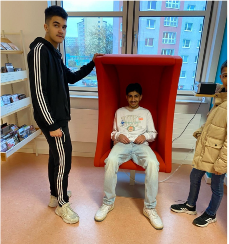 Zwei Jugendliche in einem Raum, einer sitzt in einer roten akustischen Sitzkabine, während der andere daneben steht und die Kabine festhält. Im Hintergrund sind Bücherregale und Fenster zu sehen.