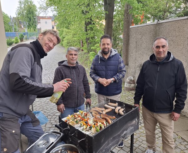 Vier Männer stehen im Freien an einem Grill mit Würstchen und Gemüsespießen.