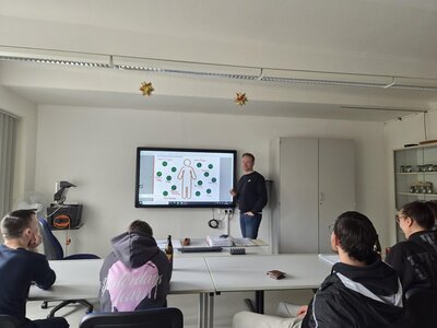 Ein Mann hält in einem Klassenzimmer einen Vortrag. Er steht neben einem Bildschirm, auf dem eine menschliche Figur mit Symbolen zu sehen ist. Fünf Auszubildende sitzen um einen Tisch und hören aufmerksam zu. Der Raum ist schlicht eingerichtet: weiße Wände, ein Schrank und ein Regal mit verschiedenen Gegenständen. Von der Decke hängen dekorative, sternförmige Ornamente.