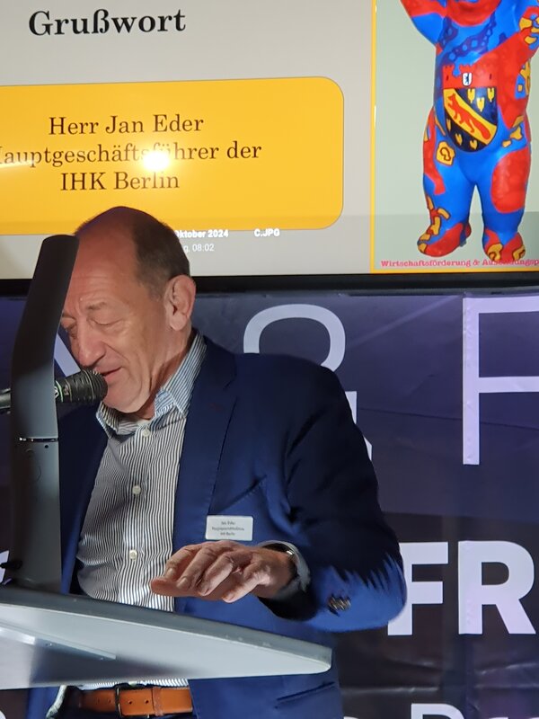 Ein Bühne mit grauem Rednerpult, an dem ein älterer Mann steht, und ein Bildschirm im Hintergrund mit der Aufschrift Ausbildungsbuddy 2024.