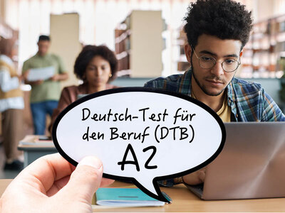Ein Mann mit Brille und lockigem Haar sitzt an einem Tisch und arbeitet an einem Laptop. Vor ihm schwebt eine Sprechblase mit dem Text 'Deutsch-Test für den Beruf (DTB) A2'. Im Hintergrund sind zwei weitere Personen, die miteinander sprechen, in einem Klassenzimmer oder Büro zu sehen.