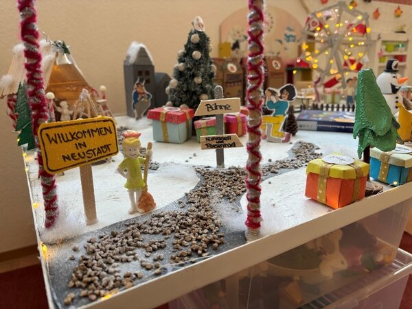 Detail einer Miniatur-Winterstadt: Schild „Willkommen in Neustadt“, Schneeweg, bunte Geschenke, kleine Figuren und ein geschmückter Baum.
