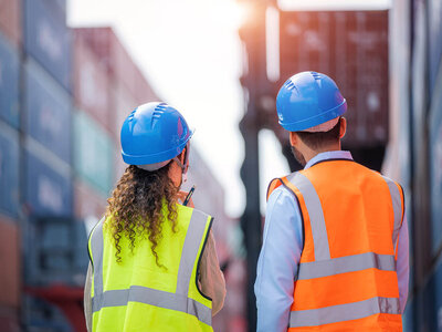 Zwei Personen stehen von hinten gesehen in einer industriellen Umgebung, vermutlich einem Container- oder Zollgelände. Beide tragen Sicherheitsausrüstung: blaue Schutzhelme und Warnwesten – eine Person in einer leuchtend gelben Weste über einem weißen Hemd mit langen, lockigen Haaren, die andere in einer orangefarbenen Weste über einem hellblauen Hemd mit kurzen Haaren. Im Hintergrund sind große, bunte Frachtcontainer gestapelt zu erkennen. Die beiden Personen scheinen eine Inspektion oder Besprechung durchzuführen. Die Atmosphäre wirkt arbeitsorientiert, professionell und passt thematisch zu Export- und Import-Prozessen.
