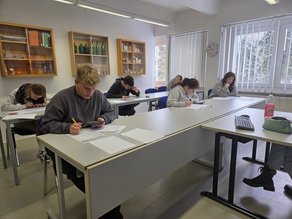Mehrere Schüler*innen sitzen in einem Klassenraum an Tischen und bearbeiten konzentriert Arbeitsblätter; vorne liegen Tastatur, Mäppchen und Getränk auf dem Tisch.