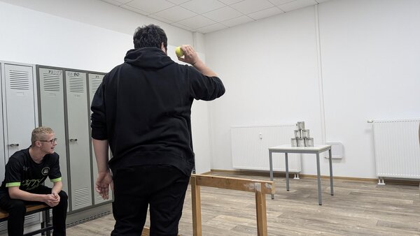 Dosenwerfen in einem Klassenraum. Ein junger Mann in schwarzen Pullover wirft einen Tennisball auf die Dosen.