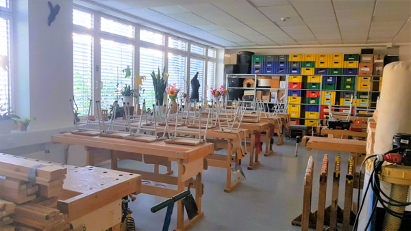 Heller Werkraum mit Reihen von Holzwerkbänken, hochgestellten Stühlen, vielen Pflanzen an den Fenstern und einer Regalwand mit bunten Kunststoffkisten für Material und Werkzeuge.