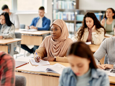 Eine Gruppe von Personen sitzt in einem Klassenzimmer oder Seminarraum. Im Vordergrund sitzt eine Frau mit einem beigen Hijab und einem beigen Pullover, die auf ein Blatt Papier schreibt. Andere Personen im Raum scheinen ebenfalls Notizen zu machen oder zuzuhören. Der Raum ist mit Tischen und Stühlen ausgestattet, und im Hintergrund sind Bücherregale zu sehen. Die Atmosphäre wirkt konzentriert und lernorientiert.