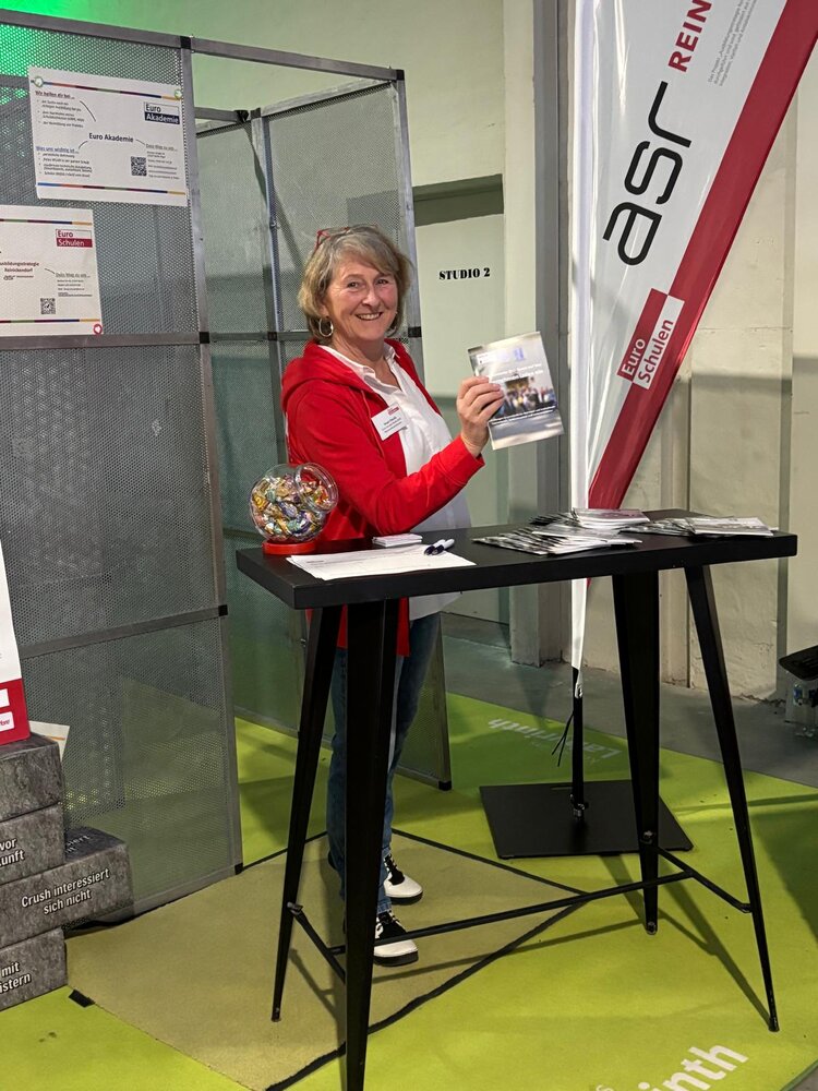 Mitarbeiterin am Infostand der Euro-Schulen: Sie lächelt in die Kamera und hält ein Informationsheft hoch; auf dem Stehtisch liegen Flyer und ein Glas mit Give-aways, daneben steht ein Euro-Schulen-Banner.