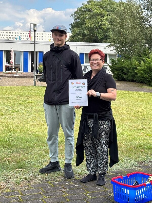 Ein junger Auszubildender und die Schulleitung stehen auf einer Wiese und halten gemeinsam eine Urkunde für den 1. Platz beim Sportfest in die Kamera.