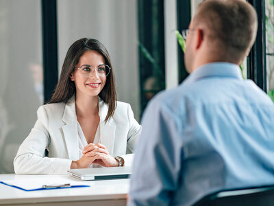 Eine Frau mit langen dunklen Haaren und einem weißen Blazer sitzt an einem Tisch und lächelt. Sie hat die Hände gefaltet und scheint sich in einem Gespräch oder einer Besprechung mit einer anderen Person zu befinden, deren Rücken zu sehen ist. Auf dem Tisch liegen ein Laptop und ein Notizbuch. Der Raum ist hell und hat große Fenster, die Tageslicht hereinlassen.