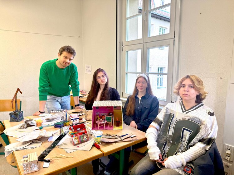 Drei Schülerinnen und ein Lehrer sitzen und stehen an einem Tisch mit Bastelmaterialien und einem kleinen Modellhaus in einem hellen Raum. Sie wirken konzentriert beim gemeinsamen Arbeiten an einem Projekt.