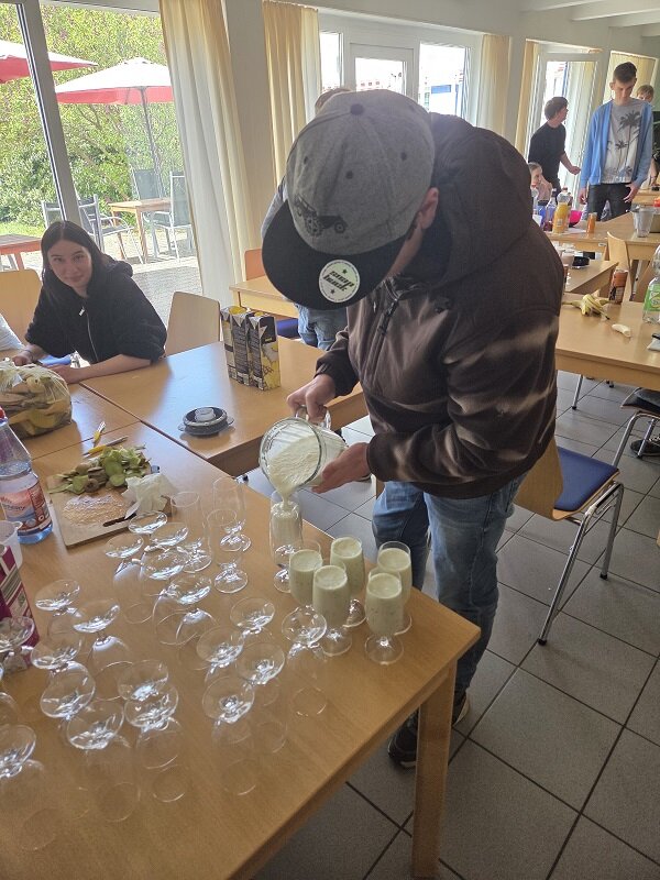 Eine Person in einer Jacke und Basecap gießt ein dickflüssiges Getränk aus einem Messbecher in ein Glas. Auf dem Tisch stehen zahlreiche leere Gläser, die für die Zubereitung von Getränken vorbereitet sind. Im Hintergrund sind weitere Personen in einem hellen Raum mit großen Fenstern zu sehen.