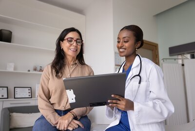 Eine Ärztin im weißen Kittel mit Stethoskop bespricht gemeinsam mit einer Patientin Unterlagen auf einem Klemmbrett. Beide sitzen in einem hellen Behandlungsraum und lächeln.