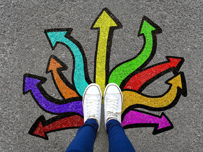 Eine Person steht auf einer Straße mit bunten, gemalten Pfeilen in verschiedenen Richtungen. Die Füße der Person, die weiße Turnschuhe und blaue Hosen trägt, sind im Vordergrund zu sehen. Die Pfeile sind in verschiedenen Farben wie Gelb, Grün, Blau, Lila, Orange und Rot gemalt und zeigen in unterschiedliche Richtungen, was eine Entscheidungssituation oder verschiedene Wege symbolisieren könnte.