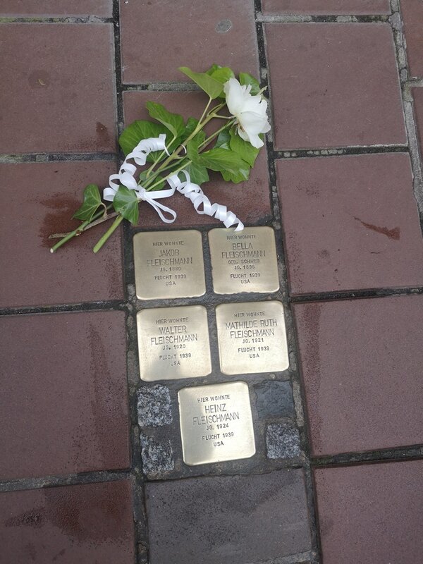Mehrere Stolpersteine zum Gedenken an die jüdische Familie Fleischmann sind in ein rotes Gehwegpflaster eingelassen. Die Messingplatten tragen die Inschriften mit den Namen Jakob Fleischmann, Bella Fleischmann, Walter Fleischmann, Mathilde Ruth Fleischmann und Heinz Fleischmann sowie Hinweise auf ihre Flucht im Jahr 1939 in die USA. Auf den Stolpersteinen liegt eine weiße Blume mit grünen Blättern und einer dekorativen weißen Schleife als Zeichen des Erinnerns und Gedenkens. Das Bild symbolisiert die Erinnerungskultur an die Opfer des Nationalsozialismus, das Schicksal jüdischer Familien während der NS-Zeit und das dezentrale Mahnmal-Projekt „Stolpersteine“ im öffentlichen Raum.