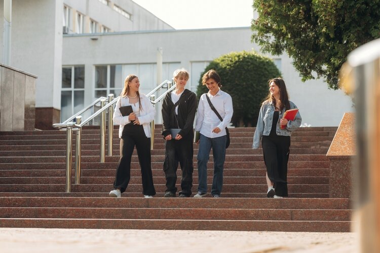 Vier junge Menschen gehen nebeneinander eine breite Treppe hinunter. Sie tragen lässige Kleidung und halten jeweils verschiedene Gegenstände in den Händen, darunter ein Notizbuch und eine Mappe. Im Hintergrund ist ein modernes, helles Gebäude mit großen Fenstern und Metallgeländern zu sehen. Rechts steht ein Baum mit dichter Krone.