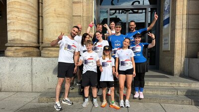 Eine Gruppe Menschen in Sporttrikots steht vor einem Sandsteingebäude. Die Menschen lächeln und strecken die Arme in die Höhe.