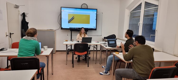 In einem Seminarraum sitzt eine Dozentin mit Laptop vor einem großen Monitor, auf dem eine Website mit gelbem Bild angezeigt wird, ihr gegenüber sitzen drei Teilnehmende an Tischen mit Laptops, Tassen und Gebäck und folgen dem Unterrichtsgespräch.