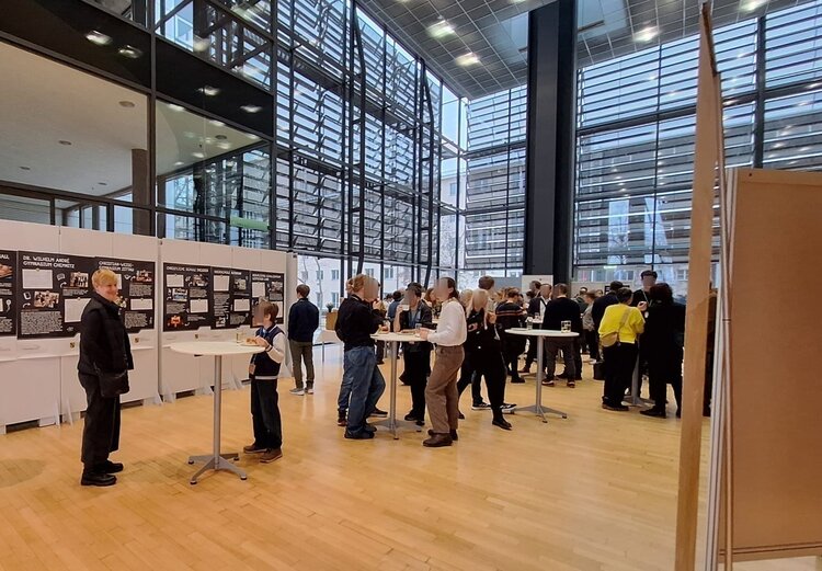 In einem hellen Foyer sind Erwachsene und junge Menschen um Stehtische versammelt, im Hintergrund hängen Infoposter an der Wand.