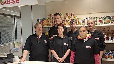 Alles neu macht der… September Drei Schülerinnen, zwei Schüler und eine Lehrerin stehen hinter der Theke des Lehrgeschäfts und strahlen in die Kamera. Sie tragen über ihren eigentlichen Kleidungstücken Polohemden als Arbeitsschutzbekleidung. In den Regalen hinter den Personen stehen Produkte, die man aus dem Supermarkt kennt, darunter Chips, Fertigsuppen, Kaffeefilter und Capri-Sun. In der linken Ecke kann man Teile einer Kasse erkennen, darüber auch ein Schild mit der Aufschrift "KASSE".
