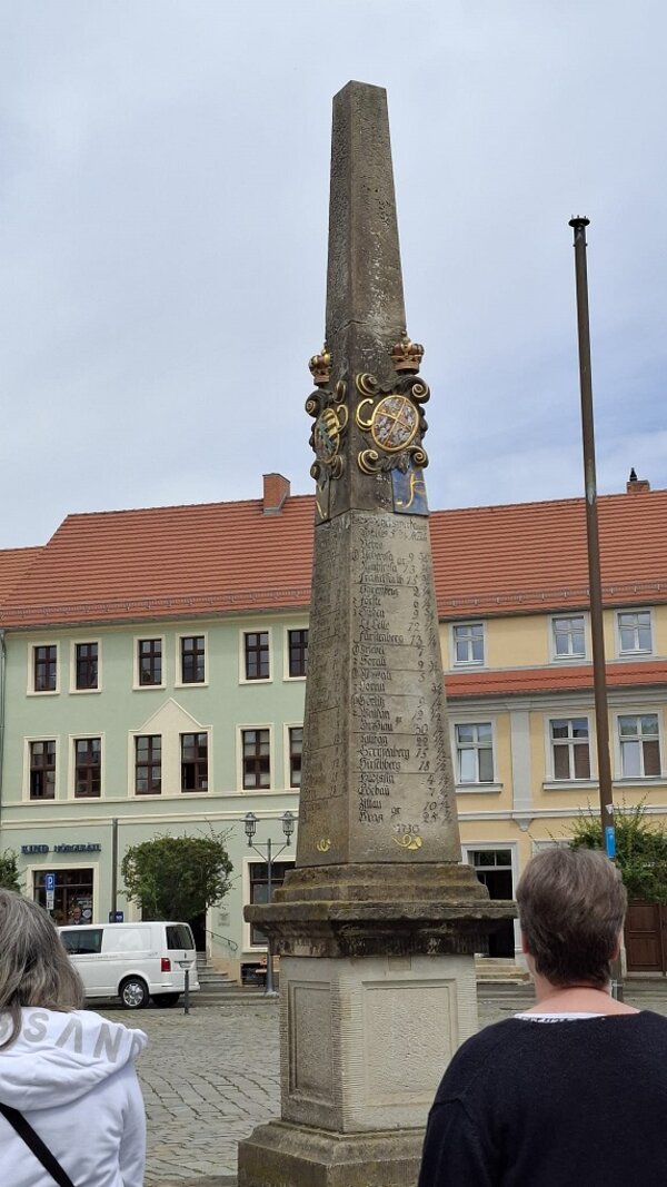 Eine alte Postsäule aus Stein mit Inschriften und goldener Verzierung mitten auf einem Platz; im Vordergrund stehen zwei Menschen. Im Hintergrund sind Häuser und ein Auto zu sehen.