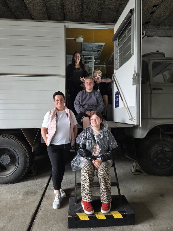 Fünf junge Menschen posieren vor einem weißen LKW mit offener Tür in einer Halle. Zwei Personen sitzen auf der Treppe zum Fahrzeug, eine Person sitzt vorne auf einem Hocker und trägt einen durchsichtigen Regencape. Zwei weitere Personen stehen seitlich und lächeln in die Kamera.