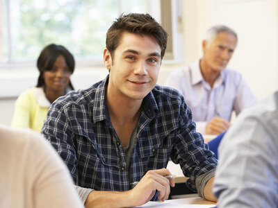 Ein junger Mann mit einem karierten Hemd sitzt an einem Tisch in einem Klassenzimmer oder Seminarraum und hält einen Stift in der Hand. Er lächelt und scheint aufmerksam zu sein. Im Hintergrund sind weitere Personen zu sehen, darunter eine Frau mit kurzem Haar und einem gelben Pullover sowie ein älterer Mann mit grauem Haar und einem lila Hemd. Der Raum ist hell und hat große Fenster, die Tageslicht hereinlassen.