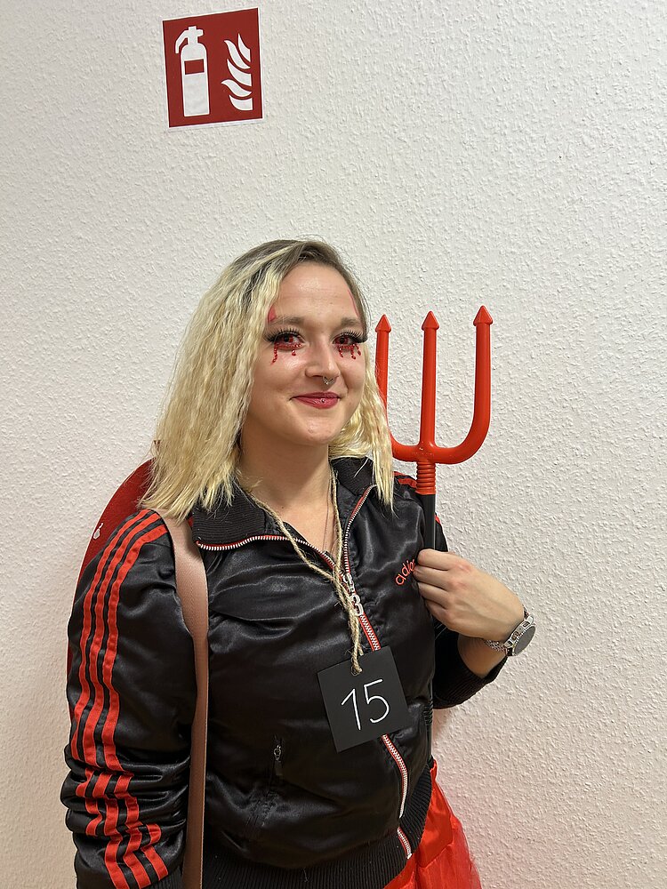 Porträt einer Teilnehmerin im Teufels-Kostüm mit Dreizack und Startnummer 15 vor einer weißen Wand.
