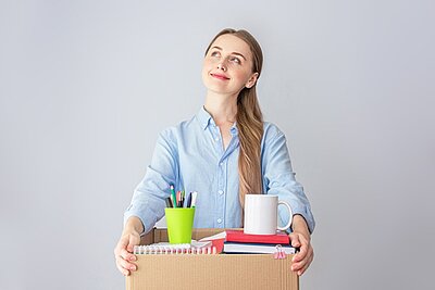 #Neustart Eine junge Frau mit blonden Haaren, die zu einem Zopf nach hinten gebunden sind, schaut optimistisch und lächelnd nach hoben und hält einen geöffneten Karton mit Schreibwaren in den Händen.