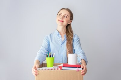 Eine junge Frau mit blonden Haaren, die zu einem Zopf nach hinten gebunden sind, schaut optimistisch und lächelnd nach hoben und hält einen geöffneten Karton mit Schreibwaren in den Händen.