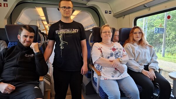 Vier Mitglieder der Pflegefachklasse sitzen und stehen im modernen Intercity-Zug; alle blicken zur Kamera, bereit für einen spannenden Tagesausflug.