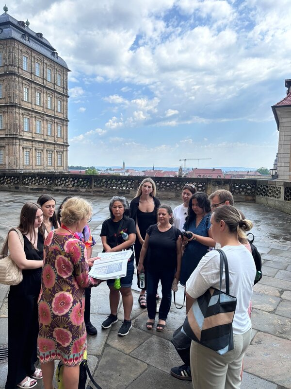 Auf einem gepflasterten Platz vor einem historischen Gebäude mit großen Fenstern und Steinbalustrade stehen die Teilnehmenden beisammen. Im Vordergrund hält eine Frau mit gemustertem Kleid ein aufgeschlagenes Heft oder Buch und spricht zu den anderen. Die Frauen hören aufmerksam zu, einige halten Taschen oder Flaschen. Der Himmel ist bewölkt, der Boden wirkt feucht.