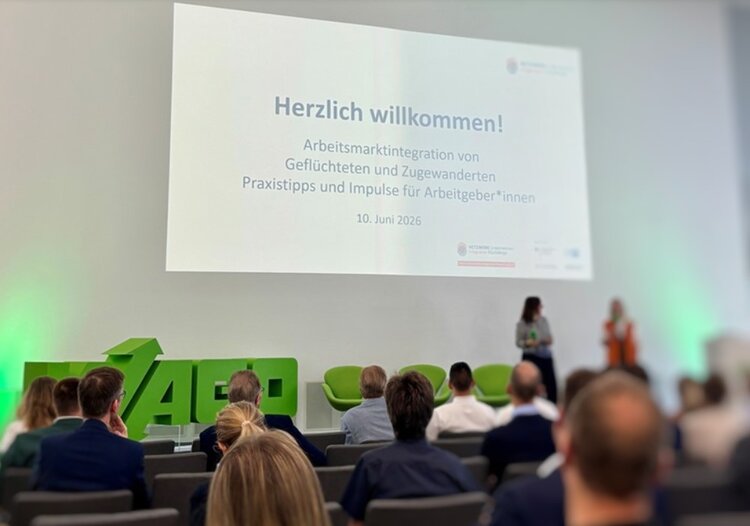 Mehrere Personen sitzen in einem modernen Konferenzraum mit weißen Wänden und grünen Sitzmöbeln. Im Vordergrund sind die Rücken von Teilnehmenden zu sehen, im Hintergrund stehen zwei Personen auf einer Bühne. An der Wand ist eine große Leinwand mit einer Präsentationsfolie zu sehen. Die Folie trägt die Überschrift 'Herzlich willkommen!' und informiert über eine Veranstaltung zur Arbeitsmarktintegration von Geflüchteten und Zugewanderten. Das Datum '10. Juni 2026' ist ebenfalls lesbar.