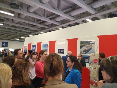 Eine Gruppe von Menschen steht vor einem Messestand mit rot-weißer Gestaltung und Infotafeln von Unternehmen wie MSC und AIDA. Eine Frau im blauen Blazer spricht zu den Anwesenden. Der Stand scheint sich auf die Kreuzfahrt- oder Tourismusbranche zu beziehen.