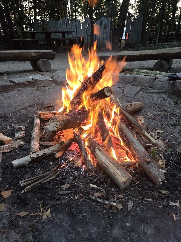 Ein hell loderndes Lagerfeuer. Im Hintergrund sind Holzbänke und Bäume in der Dämmerung zu sehen.