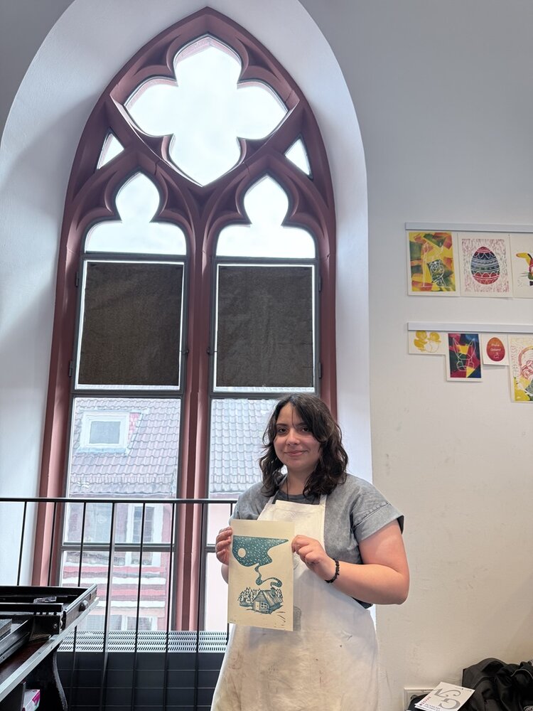 Eine junge Teilnehmerin mit schulterlangem Haar steht vor einem großen gotischen Fenster mit rotbraunem Rahmen, hält ein Blatt Papier mit einer blauen Grafik hoch und lächelt in die Kamera. Sie trägt eine helle Schürze über einem grauen Oberteil. Im Hintergrund sind mehrere farbige Zeichnungen und Grafiken an der Wand befestigt. Das Fenster zeigt vier dekorative Bögen und blickt auf ein Gebäude mit Ziegeldach.