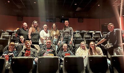 Abwechslung im Schulalltag: ein Kinobesuch Ein dunkler Kinosaal mit Sesseln. In den Reihen stehen und sitzen 13 junge Menschen und blicken lächelnd in die Kamera.