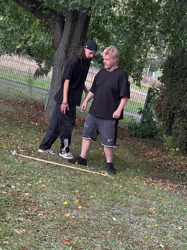 Zwei Personen stehen auf einer Wiese unter einem Baum. Beide tragen dunkle, legere Kleidung. Eine Person trägt ein schwarzes T-Shirt und kurze Hosen, die andere ein schwarzes T-Shirt und lange Trainingshose sowie eine Kopfbedeckung. Zwischen ihnen liegt ein langer Holzstab auf dem Boden. Im Hintergrund sind ein Metallzaun und weitere Bäume zu sehen.